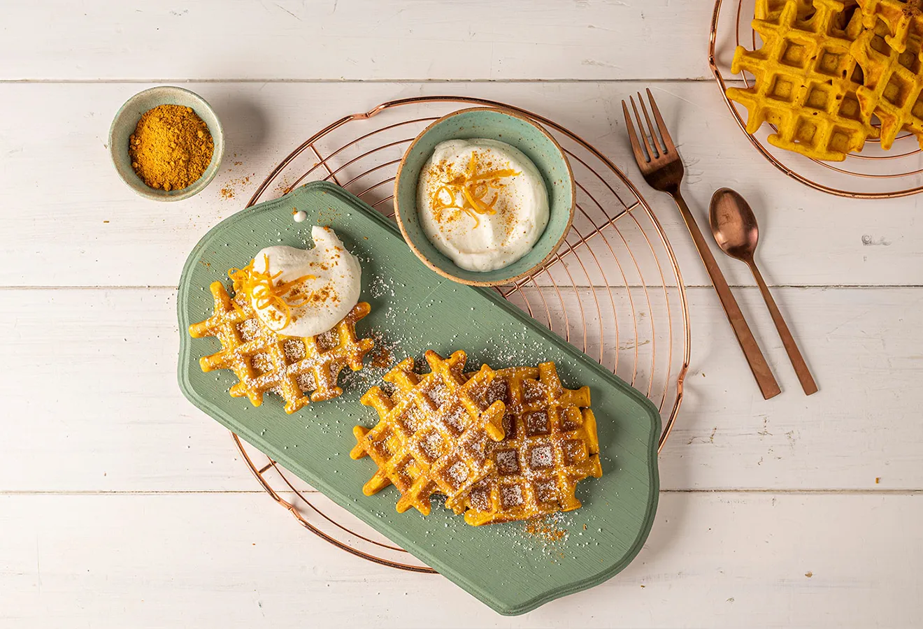 Kürbis-Waffeln mit Puderzucker und Kurkuma auf grünem Teller, serviert mit Sahne-Dip und orangefarbenen Waffeln im Hintergrund.