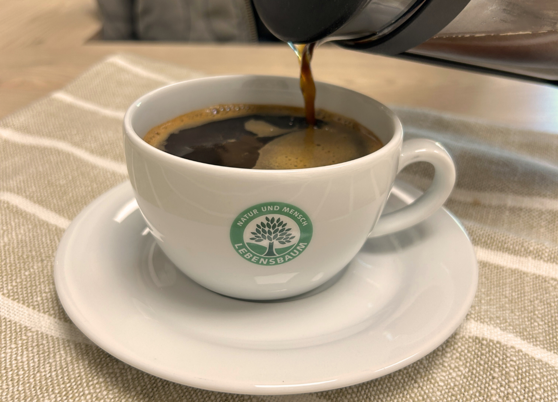 Frisch gebrühter Kaffee wird aus einer French Press in eine Tasse mit Lebensbaum-Logo gegossen