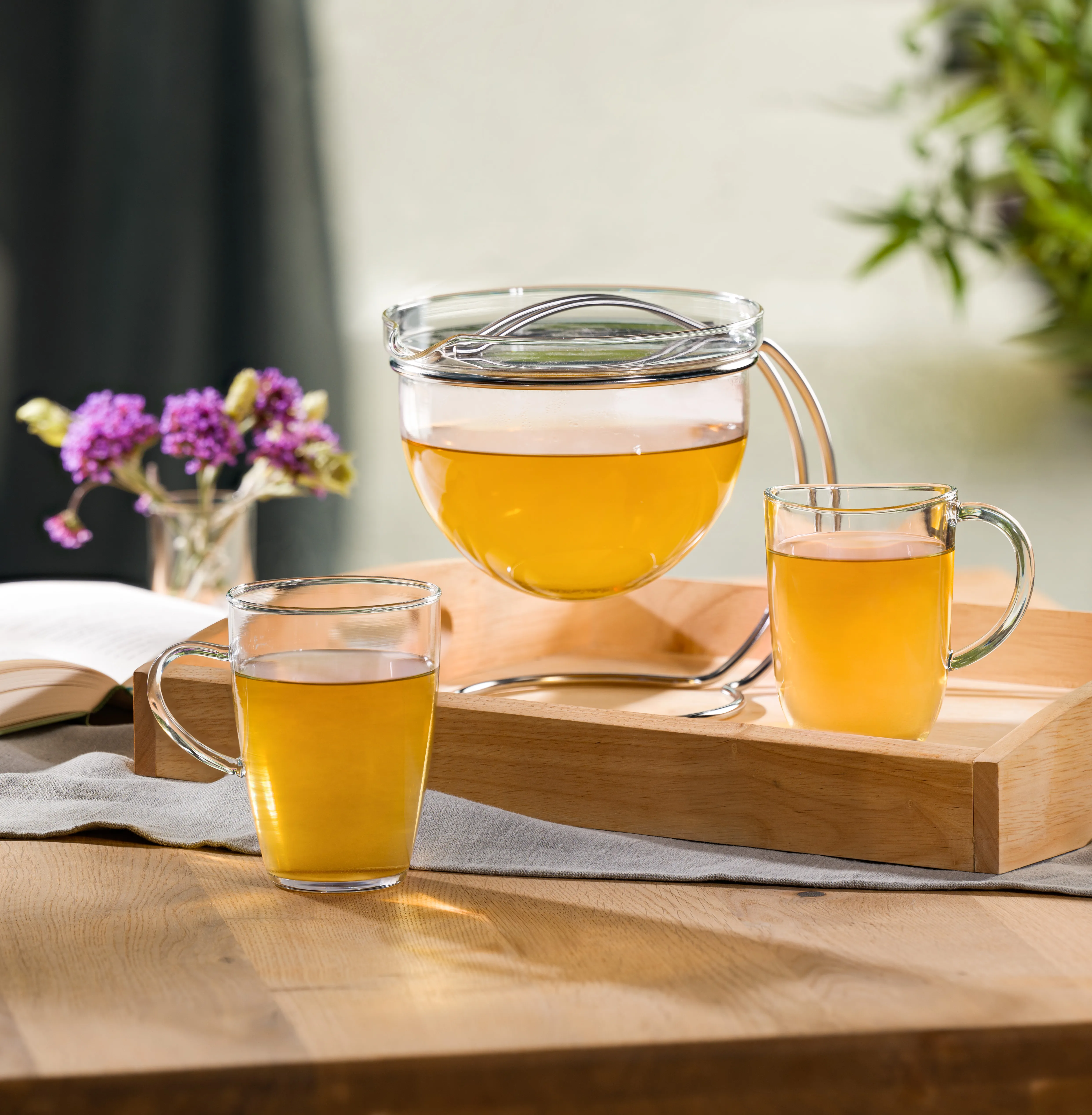 Teezubereitung mit Glaskanne und zwei Tassen goldenem Tee auf Holztablett, pink blühende Blume und aufgeschlagenes Buch im Hintergrund