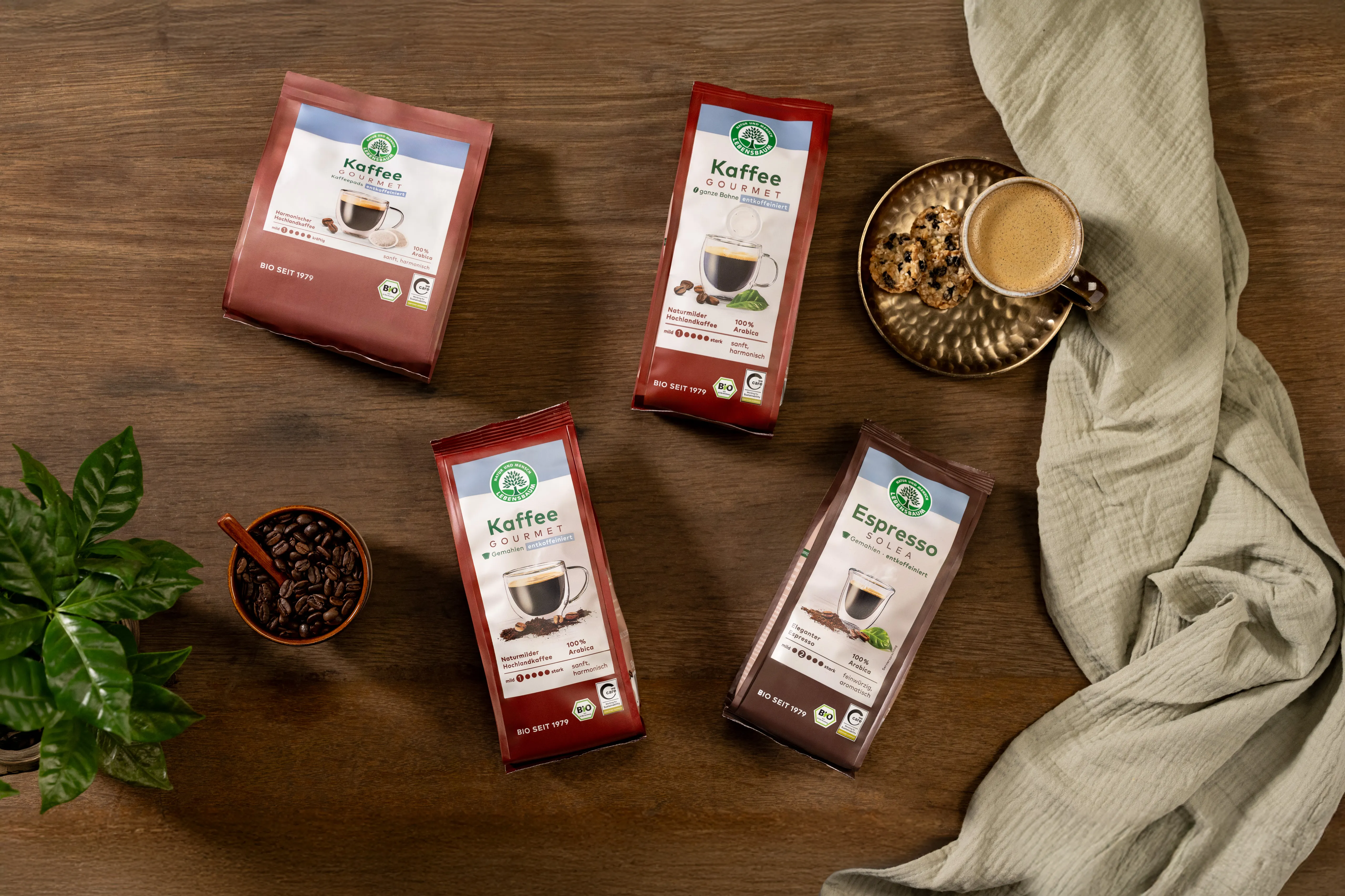 Lebensbaum Bio-Kaffee Sortiment mit roten und braunen Verpackungen auf Holztisch, daneben Kaffeetasse mit Schaum, Kekse, Kaffeebohnen in Schale und grüne Pflanze.