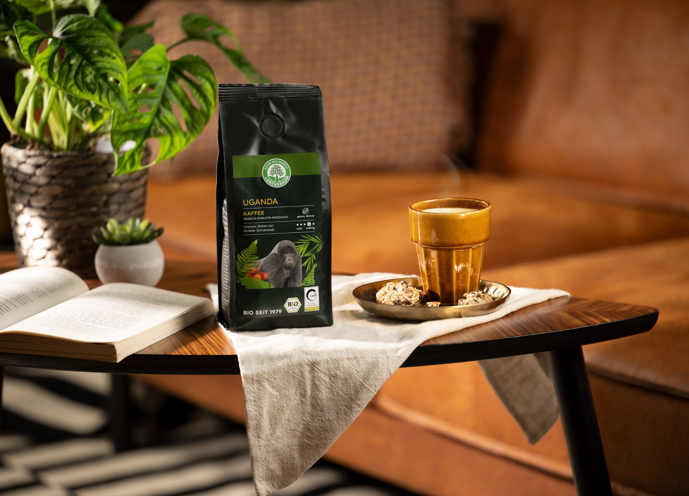 Kaffeepackung „Uganda“ mit Gorilla-Motiv steht neben einer dampfenden Tasse und Gebäck auf einem Tisch vor einem Ledersofa