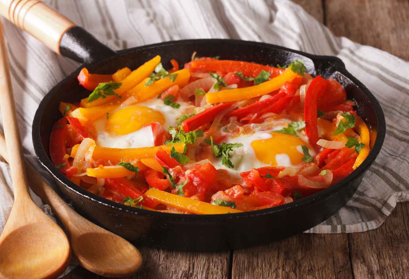 Piperade mitEi in einer schwarzen Pfanne, daneben Holzkochlöffel