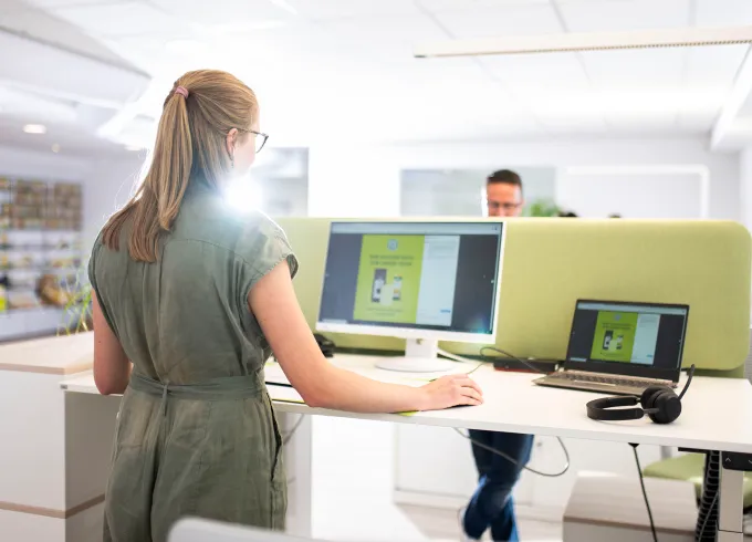 Frau mit Brille am Schreibtisch mit Monitor und Laptop in modernem Büro, grüne Trennwand, Kollege im Hintergrund.