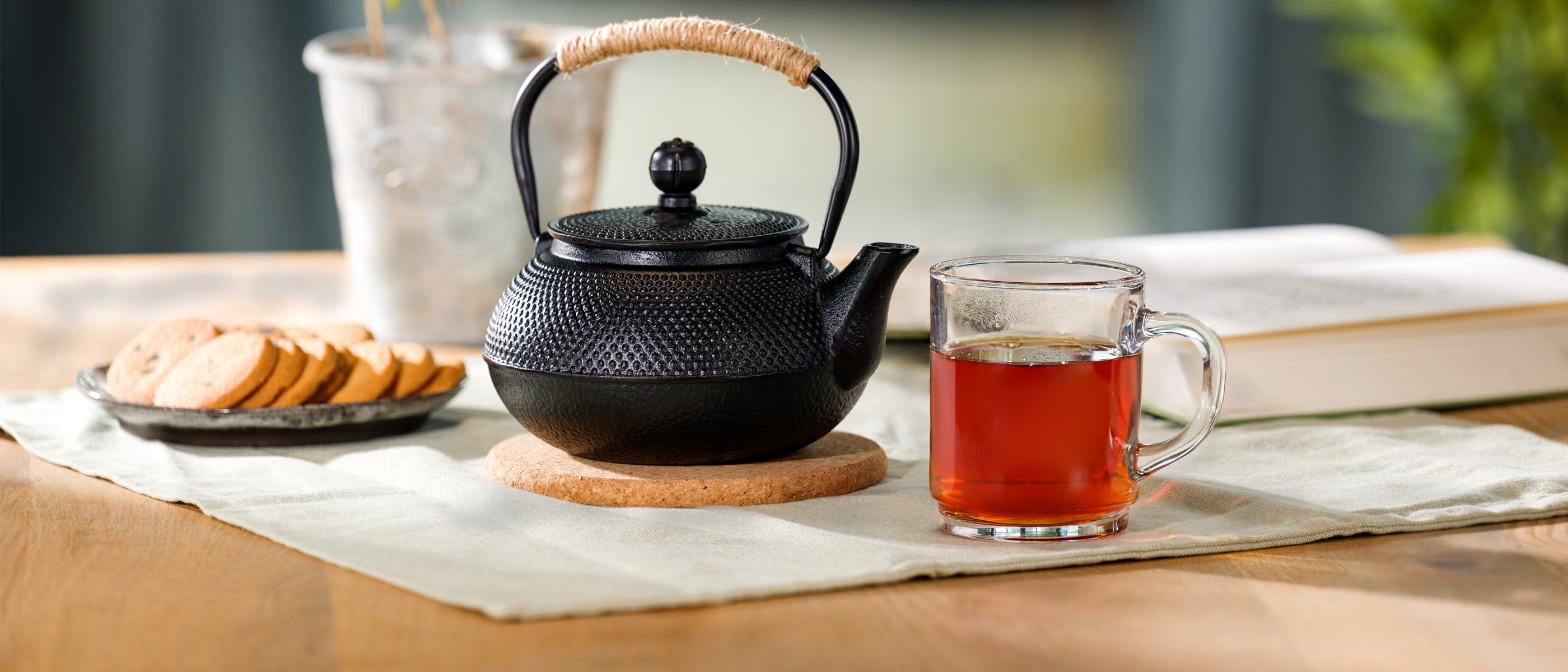 Eine schwarze Teekanne mit geflochtenem Griff steht auf einem runden Untersetzer, daneben ein Glas mit kräftigem Tee und ein Teller mit Keksen auf einem hellen Tischläufer