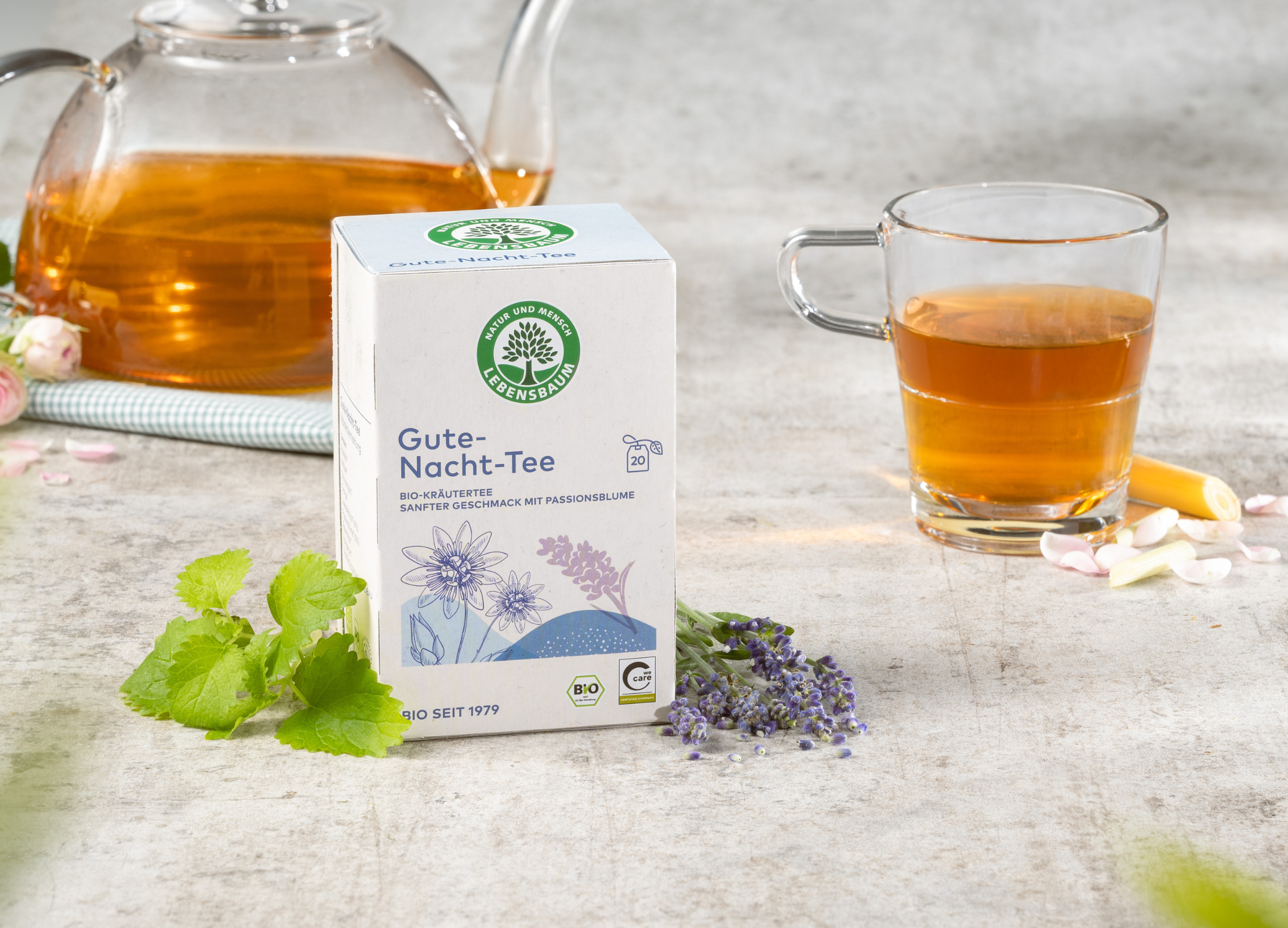 Packung Gute-Nacht-Tee von Lebensbaum mit Glas und Kanne Tee, umgeben von frischen Kräutern