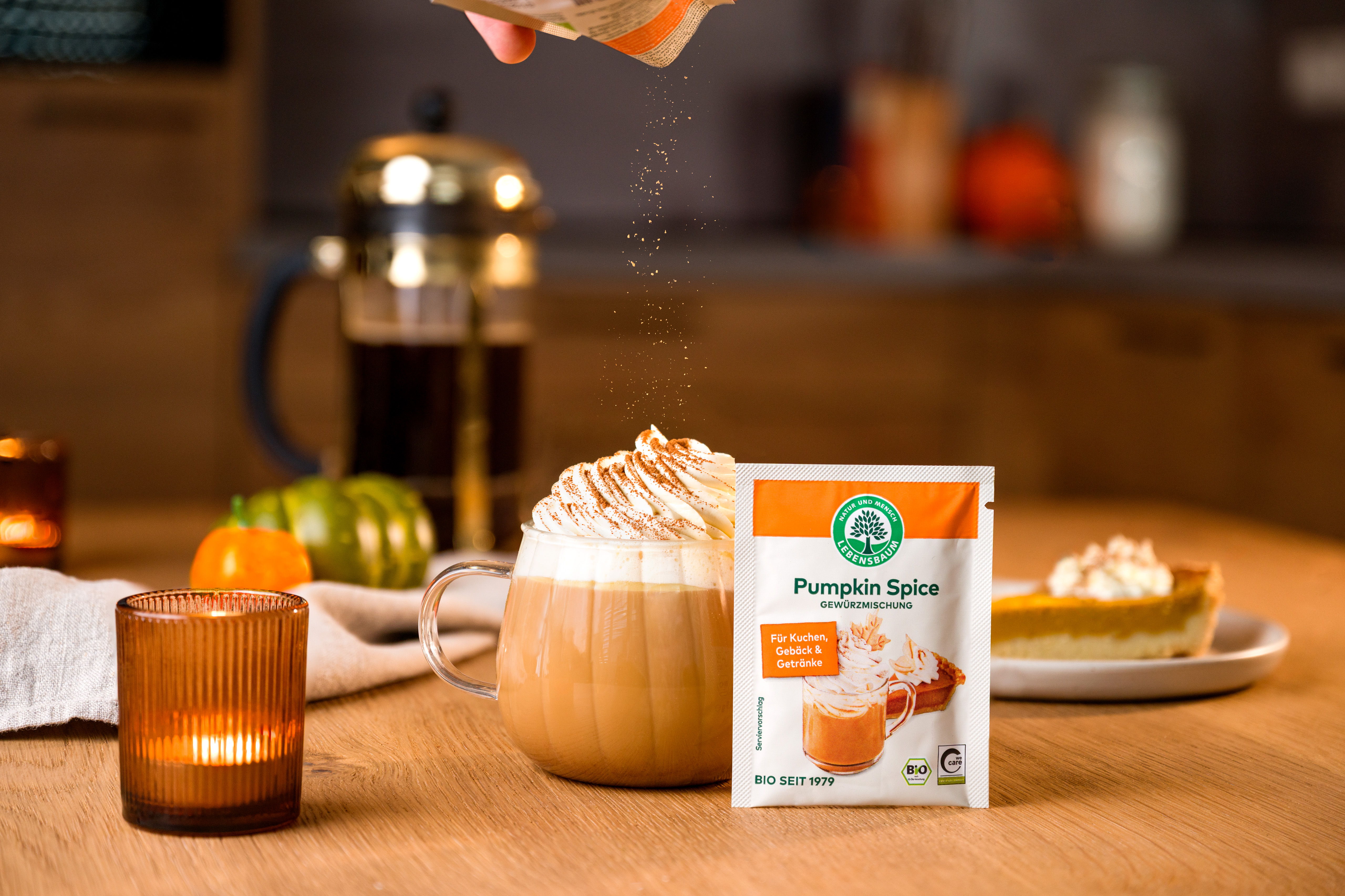 Ein Pumpkin-Spice-Gewürzpäckchen steht neben einem Glas Pumpkin Spice Latte Getränk und einem Srück Kuchen auf einem Holztisch mit herbstlicher Dekoration