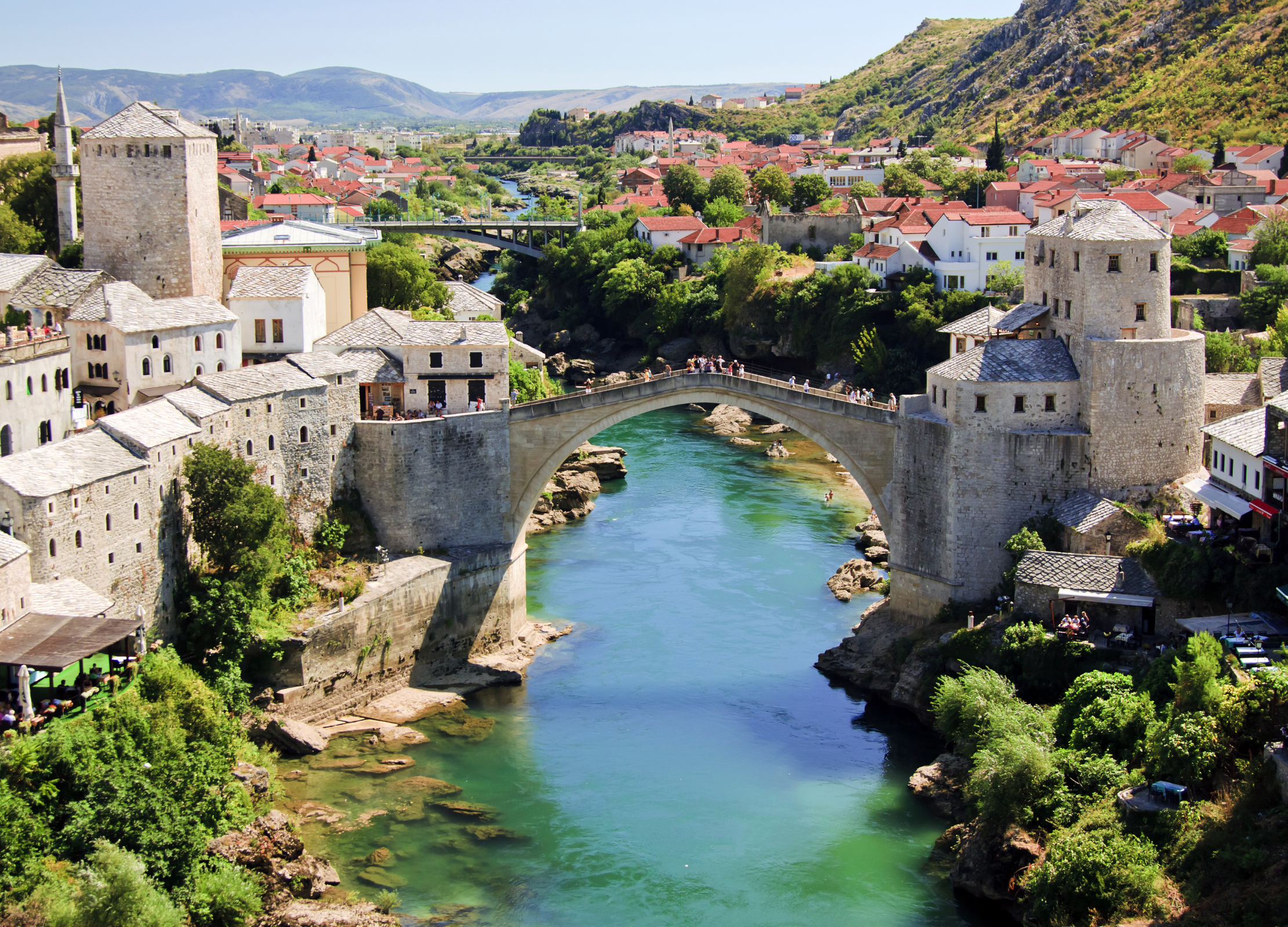 Historische Steinbrücke überspannt einen türkisfarbenen Fluss inmitten einer Stadt mit roten Dächern und Hügeln im Hintergrund in Bosnien Herzegowina