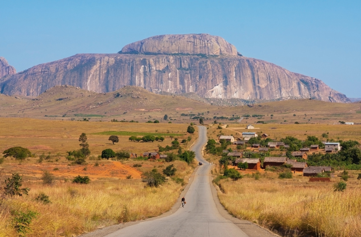 Straße führt durch Savanne auf ein Dorf zu, mit großem Tafelberg in Madagaskar im Hintergrund