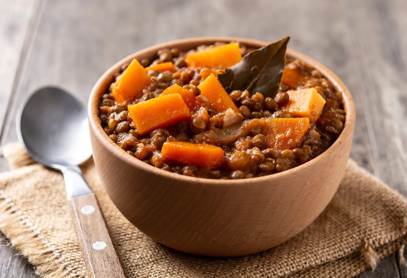 Linsen-Tajine mit Kürbis und Datteln in einem Holzschälchen mit einem Lorbeerblatt garniert, daneben ein Löffel