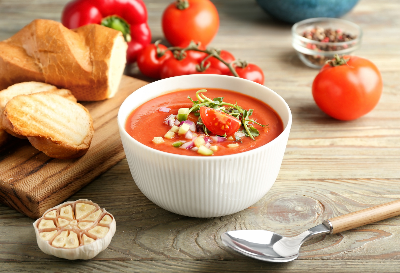 Gazpacho mit Tomate, Oregano und Zwiebelstückchen garniert in einer weißen Schale, daneben ein Löffel