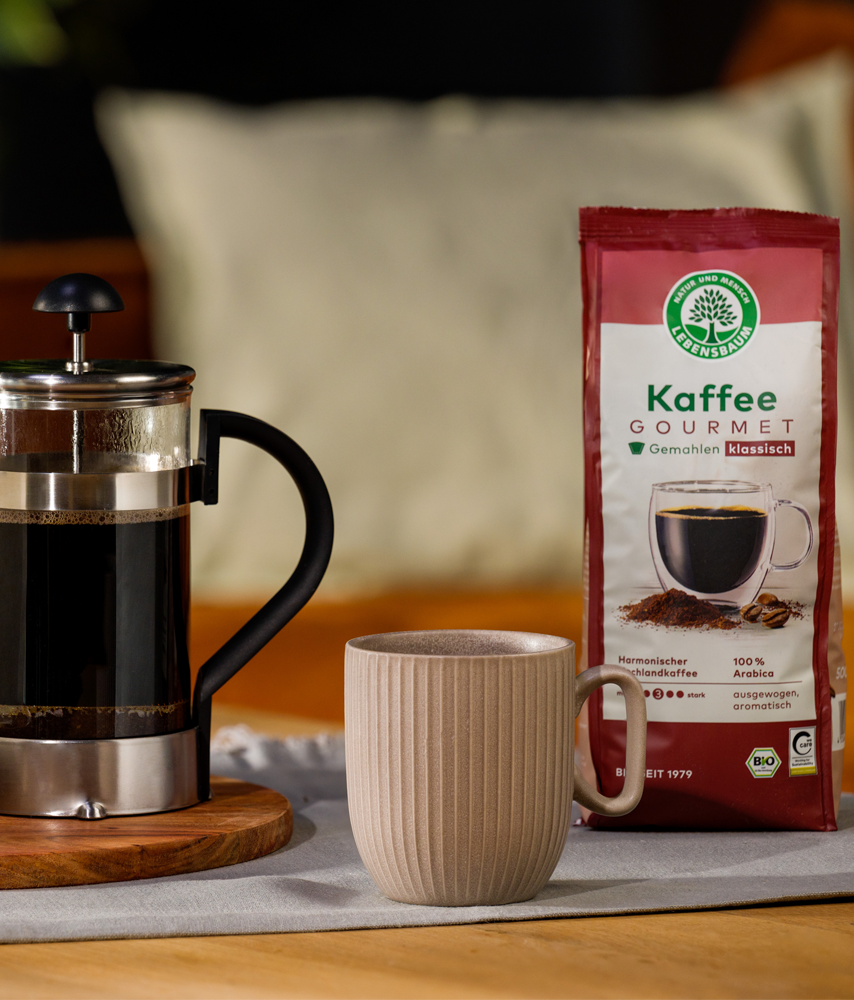 Packung Lebensbaum Kaffee Gourmet steht neben einer French Press und einer Tasse auf einem gedeckten Tisch, daneben ein kleiner Keks­teller und eine Kaffeepflanze im Topf