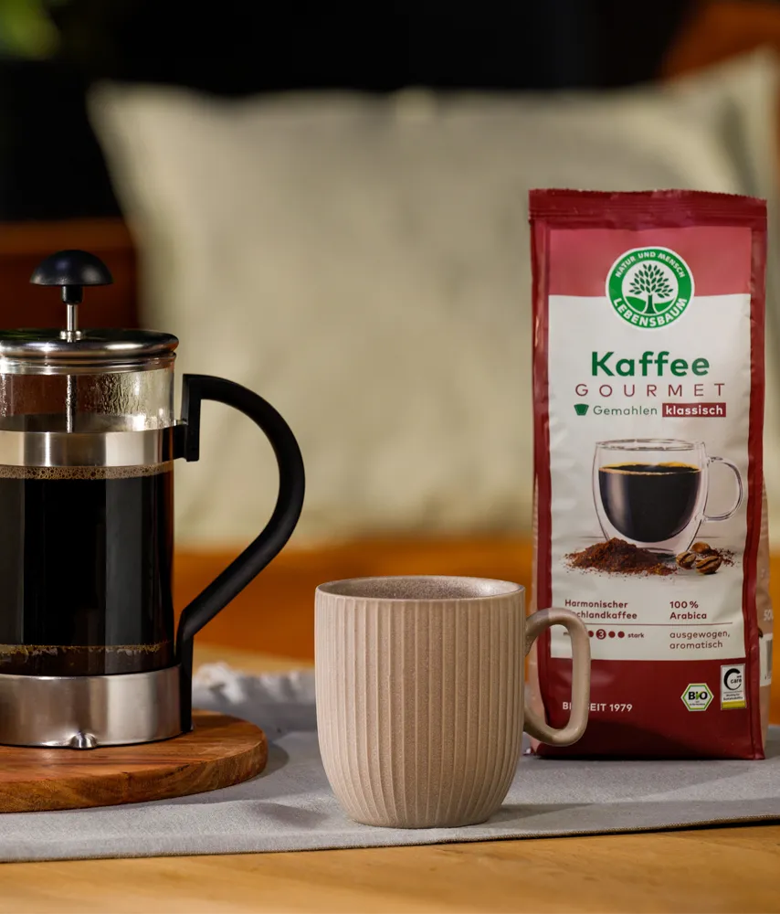 Packung Lebensbaum Kaffee Gourmet steht neben einer French Press und einer Tasse auf einem gedeckten Tisch, daneben ein kleiner Keks­teller und eine Kaffeepflanze im Topf
