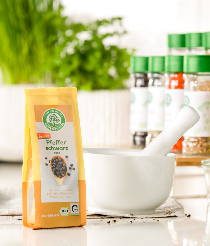Packung Lebensbaum Pfeffer schwarz steht neben einem weißen Mörser auf einer hellen Küchenarbeitsfläche, im Hintergrund frische Kräuter und Gewürze