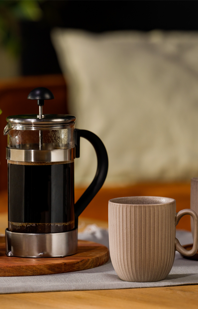 Zwei beige Tassen stehen neben einer gefüllten French Press auf einem gedeckten Tisch