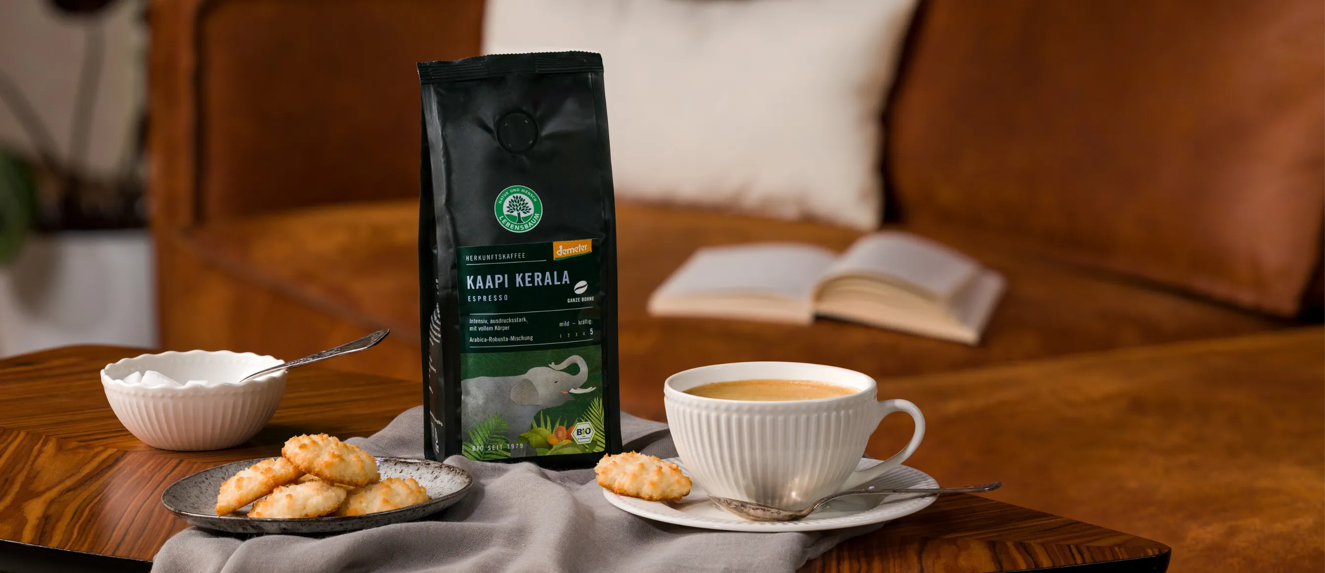 Eine Tüte unseres Kaapi Kerala auf einem Tisch, daneben eine Tasse Kaffee und ein Teller Kekse