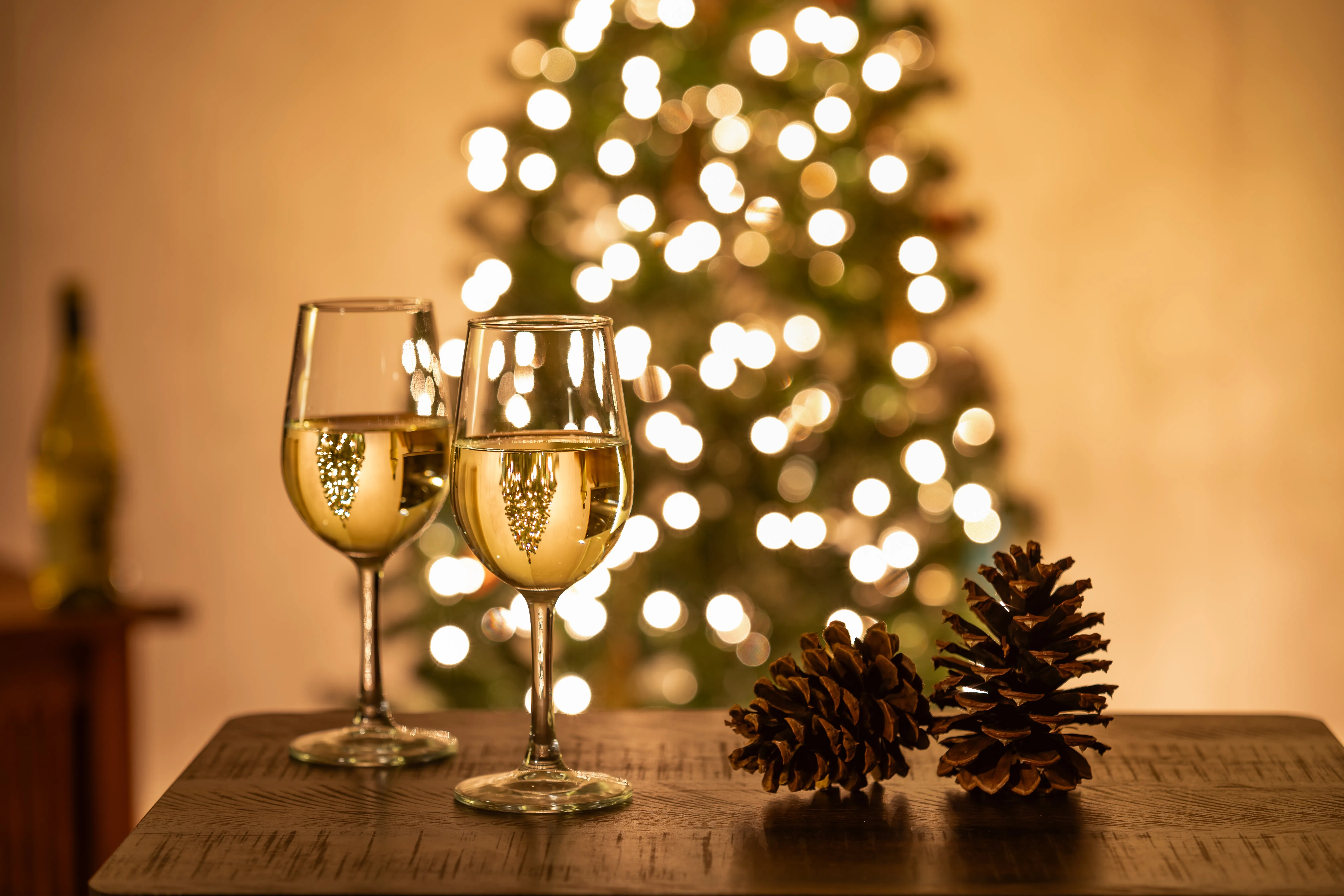 Zwei Gläser Weißwein mit Blasen vor festlich illuminiertem Weihnachtsbaum und Tannenzapfen
