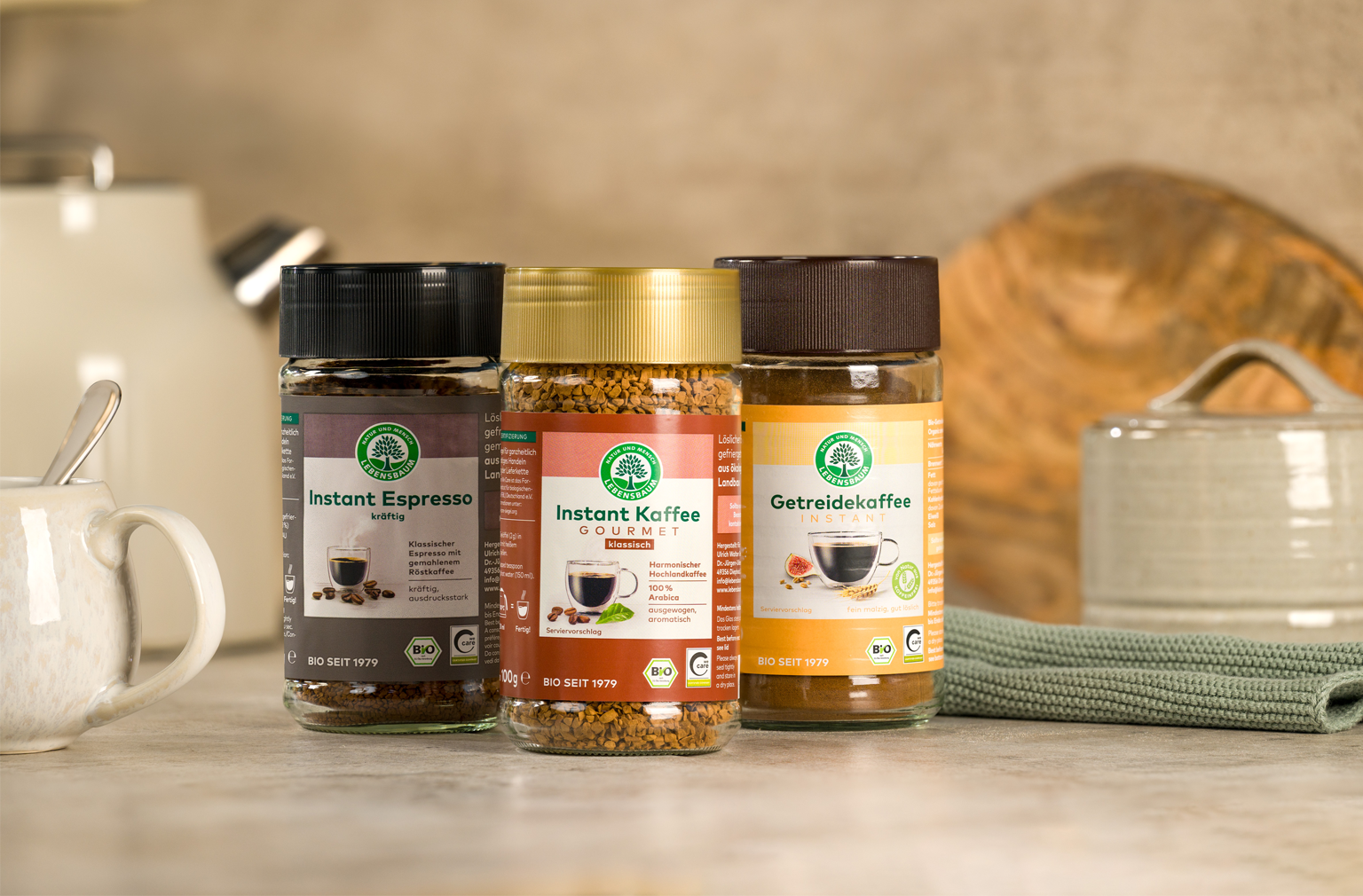 Drei Gläser Instantkaffee von Lebensbaum stehen nebeneinander auf einer Küchenarbeitsfläche, daneben eine Tasse mit Löffel und Küchendeko im Hintergrund