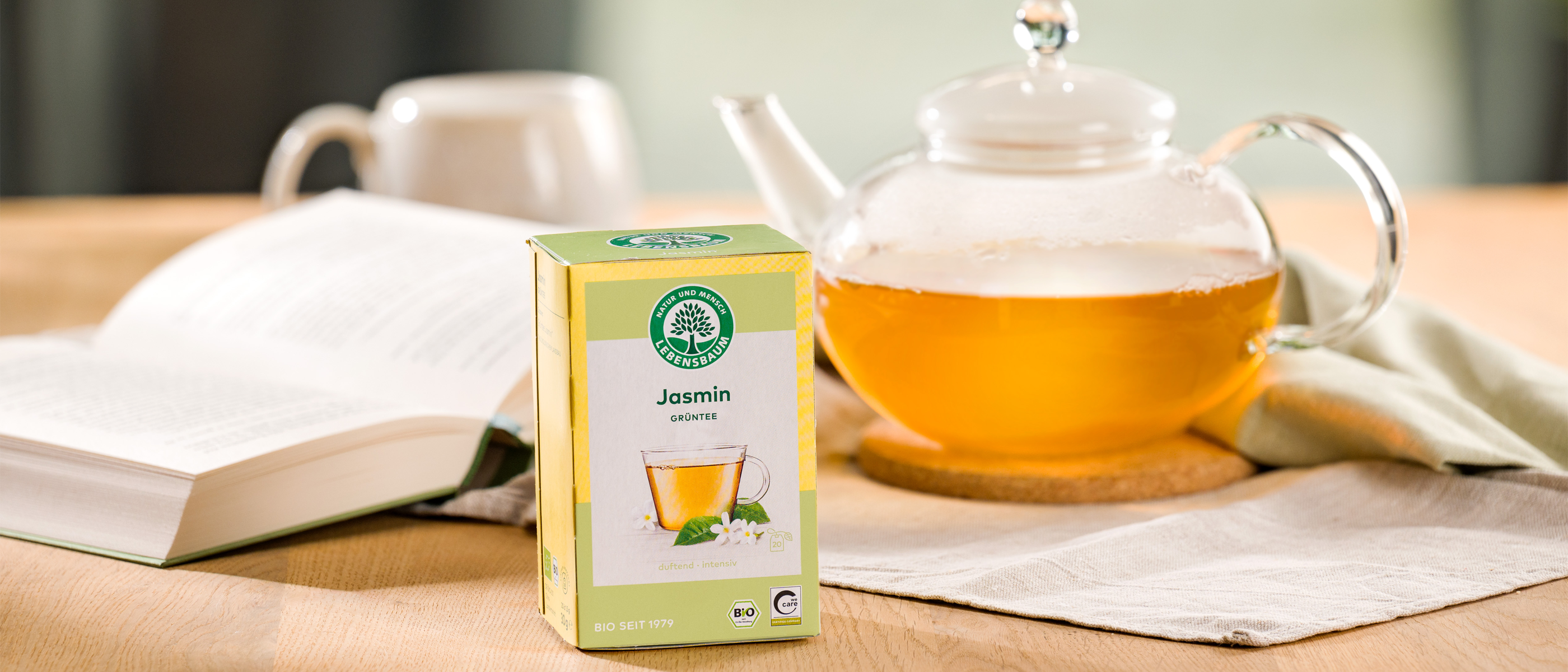 Packung Lebensbaum Jasmintee steht vor einer gläsernen Teekanne mit goldgelbem Tee – daneben ein aufgeschlagenes Buch auf einem Holztisch