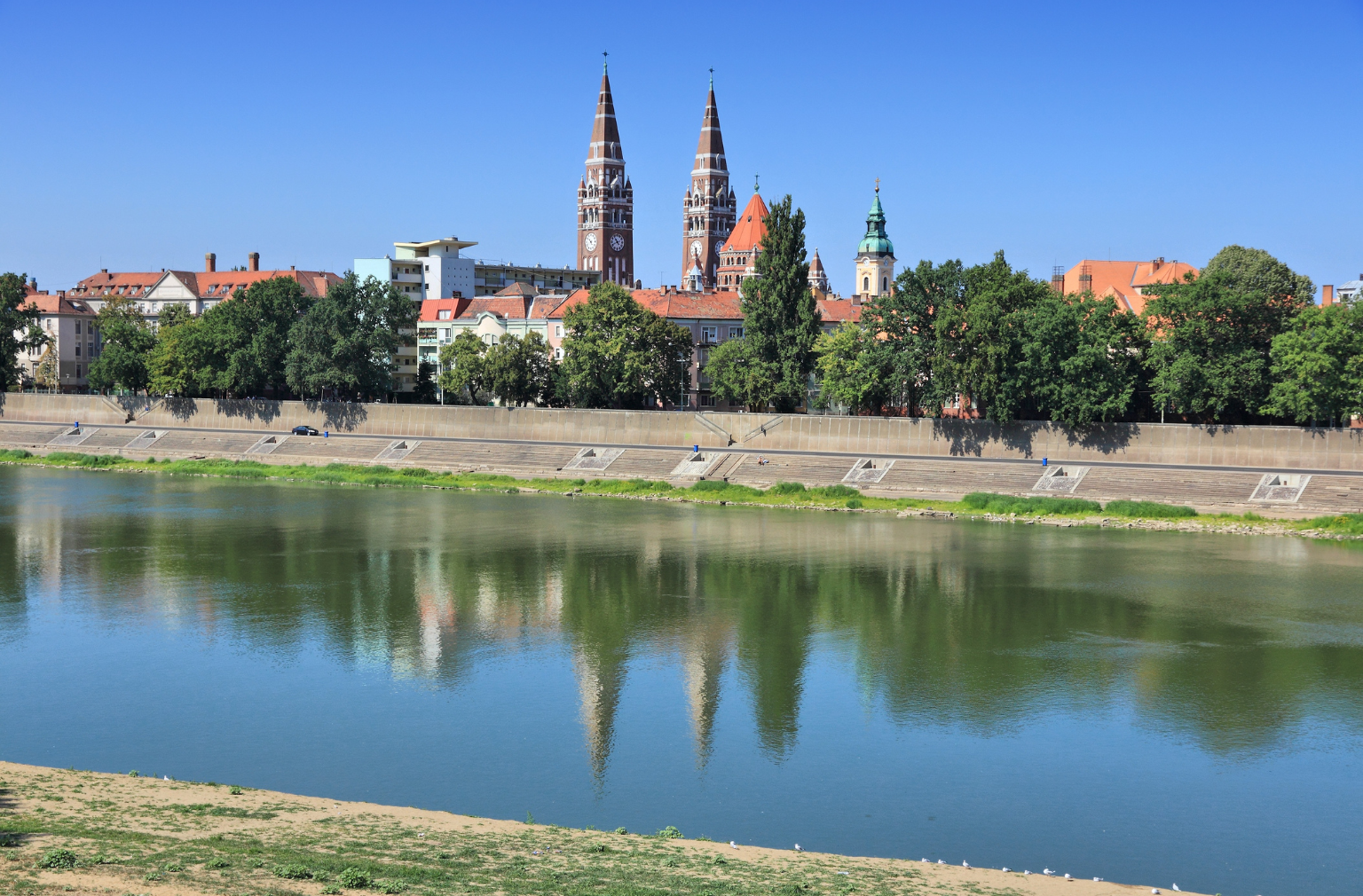 Stadtansicht mit Kirchtürmen und deren Spiegelung im Flussufer bei blauem Himmel aus Ungarn