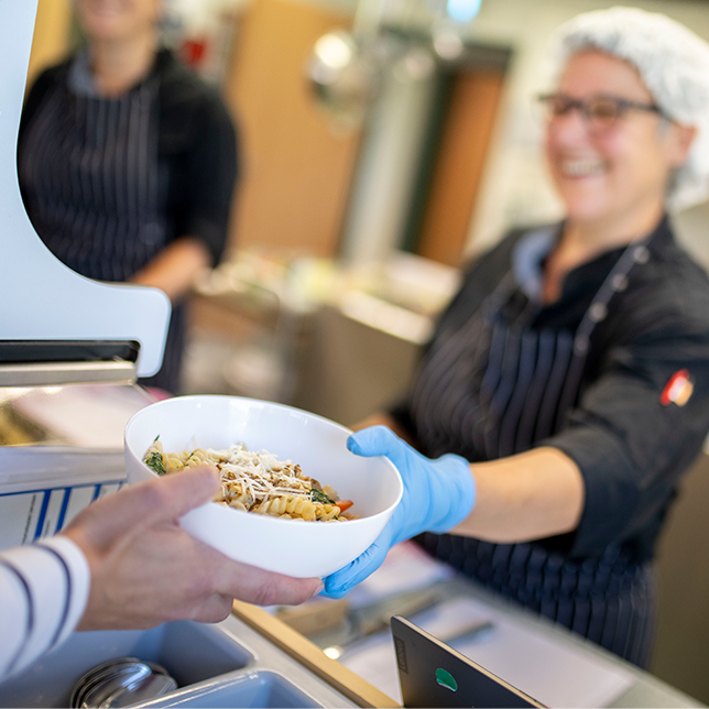 Küchenmitarbeiterin reicht eine Schale mit Pasta über die Essensausgabe