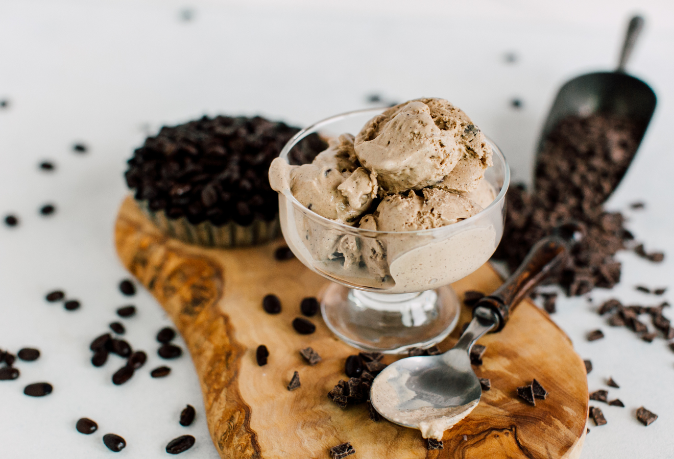 Weißes Kaffee-Eis in einem Glas-Eisbecher, daneben ein Löffel und Kaffeebohnen auf einem Holzbrett