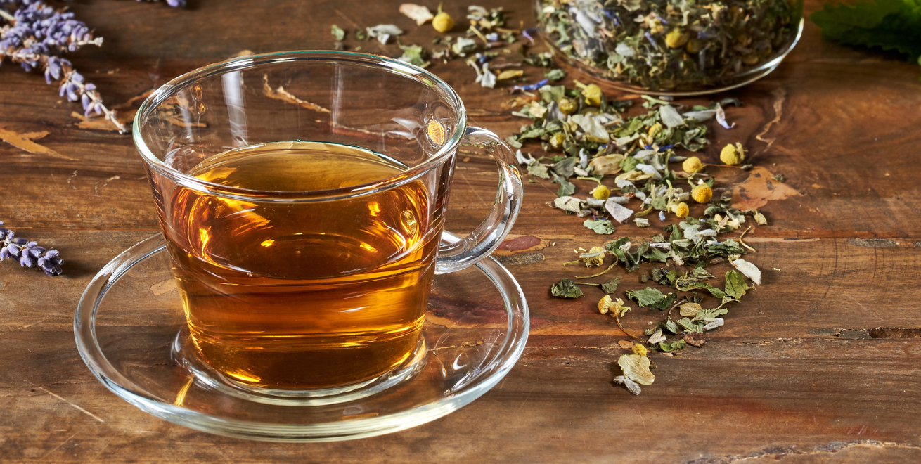 Eine Glastasse mit Kräutertee steht auf einem Holztisch. Daneben liegen ein paar frische Kräuter
