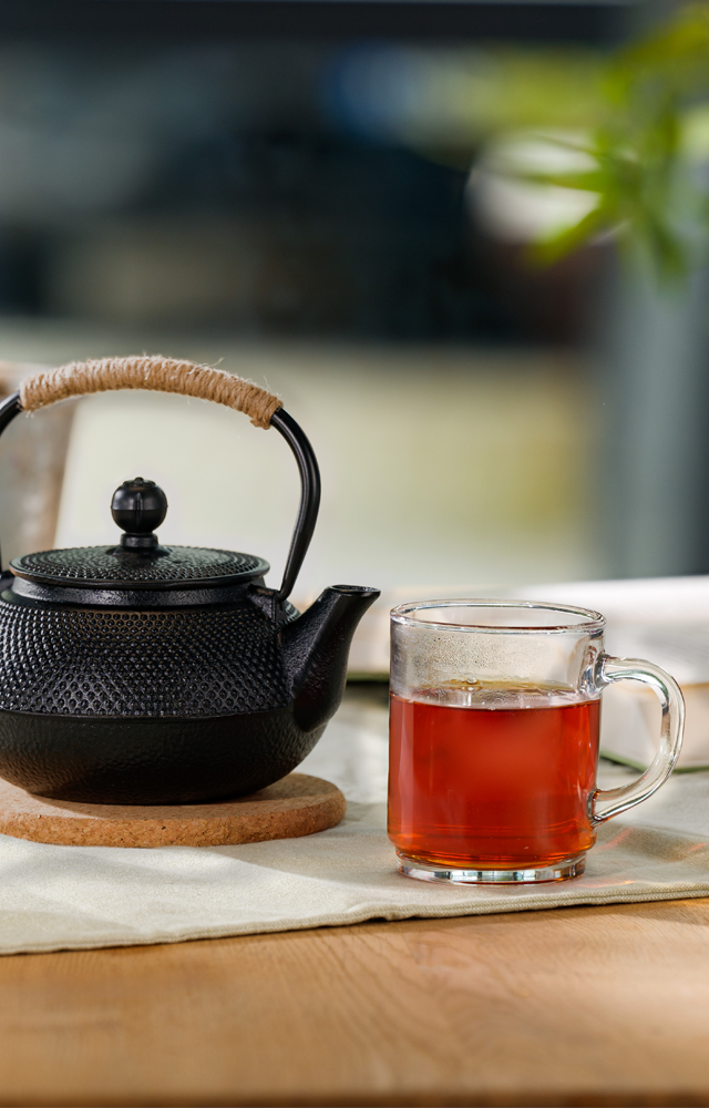 Eine schwarze Teekanne mit geflochtenem Griff steht auf einem runden Untersetzer, daneben ein Glas mit kräftigem Tee und ein Teller mit Keksen auf einem hellen Tischläufer