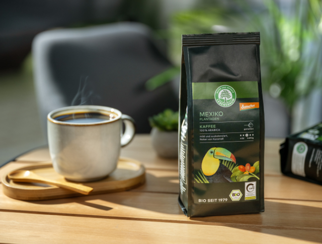 Eine Tasse dampfender Kaffee steht neben einer Packung Lebensbaum-Kaffee aus Mexiko auf einem Holztisch in sonniger Atmosphäre