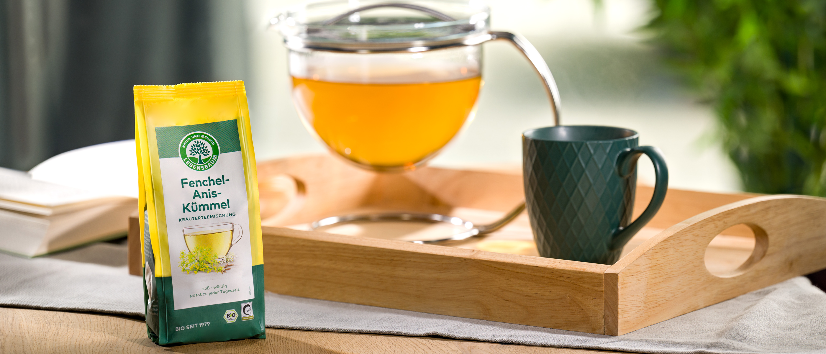 Eine Tüte Lebensbaum Fenchel-Anis-Kümmel-Tee steht neben einer gefüllten Glasteekanne und einer grünen Tasse auf einem Tablett