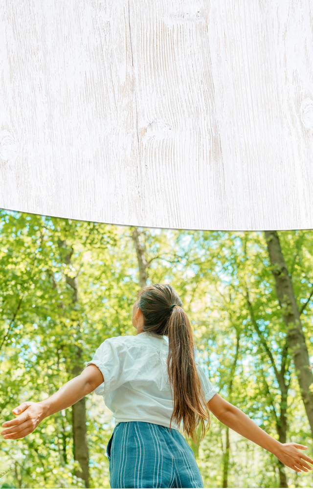 Frau mit ausgebreiteten Armen steht im sonnendurchfluteten Wald und schaut nach oben