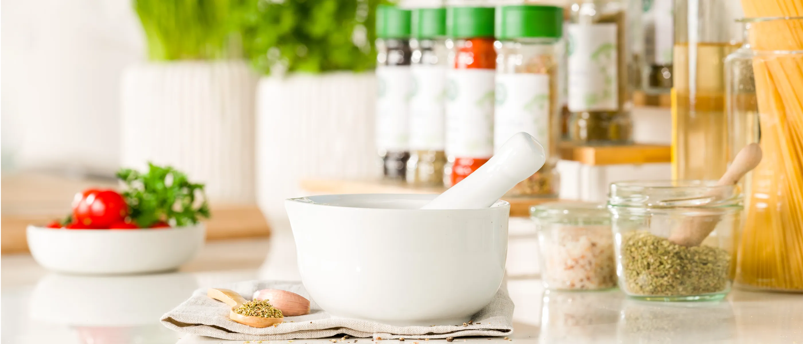 Weißer Mörser mit Stößel steht auf einem Tuch, daneben ein kleiner Löffel mit Gewürzen – im Hintergrund verschiedene Gewürzstreuer mit grünem Deckel