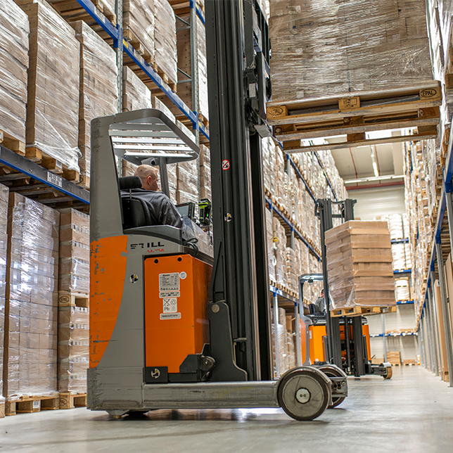 Lagermitarbeiter auf einem orangenen Gabelstapler zwischen Regalen, im Hintergrund ein weiterer Gabelstapler