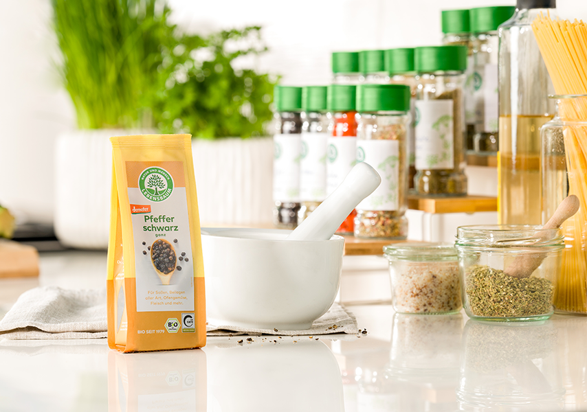Packung Lebensbaum Pfeffer schwarz steht neben einem weißen Mörser auf einer hellen Küchenarbeitsfläche, im Hintergrund frische Kräuter und Gewürze