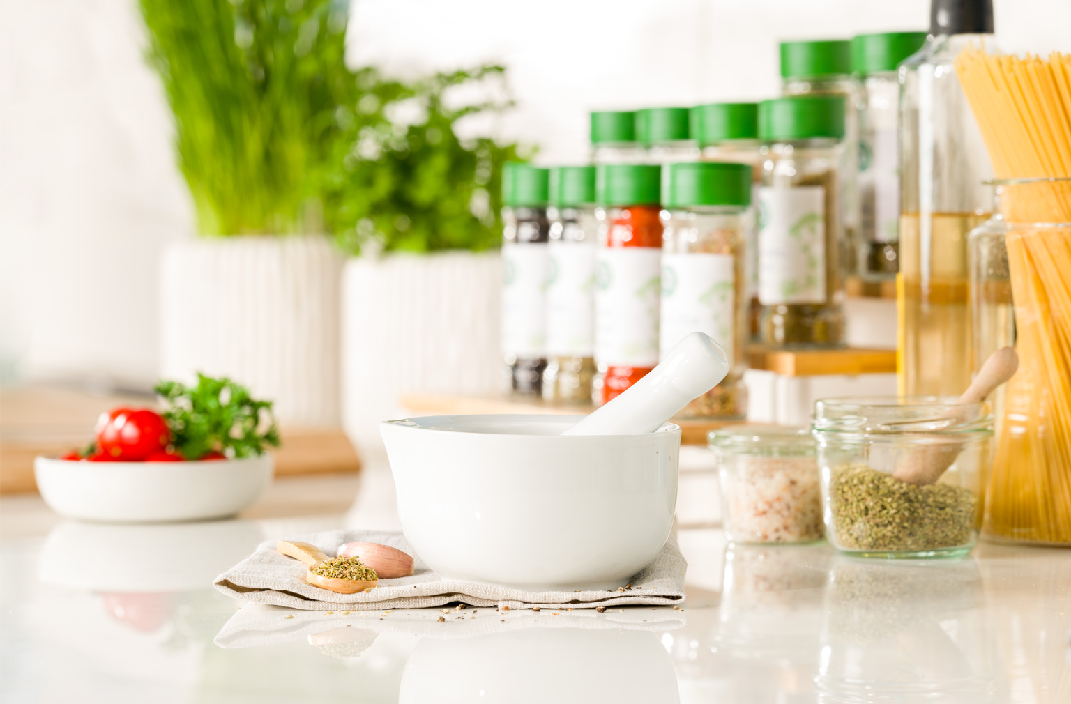 Weißer Mörser mit Stößel steht auf einem Tuch, daneben ein kleiner Löffel mit Gewürzen – im Hintergrund verschiedene Gewürzstreuer mit grünem Deckel
