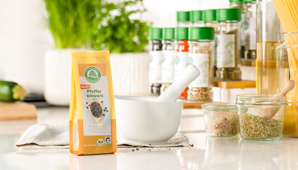 Packung Lebensbaum Pfeffer schwarz steht neben einem weißen Mörser auf einer hellen Küchenarbeitsfläche, im Hintergrund frische Kräuter und Gewürze