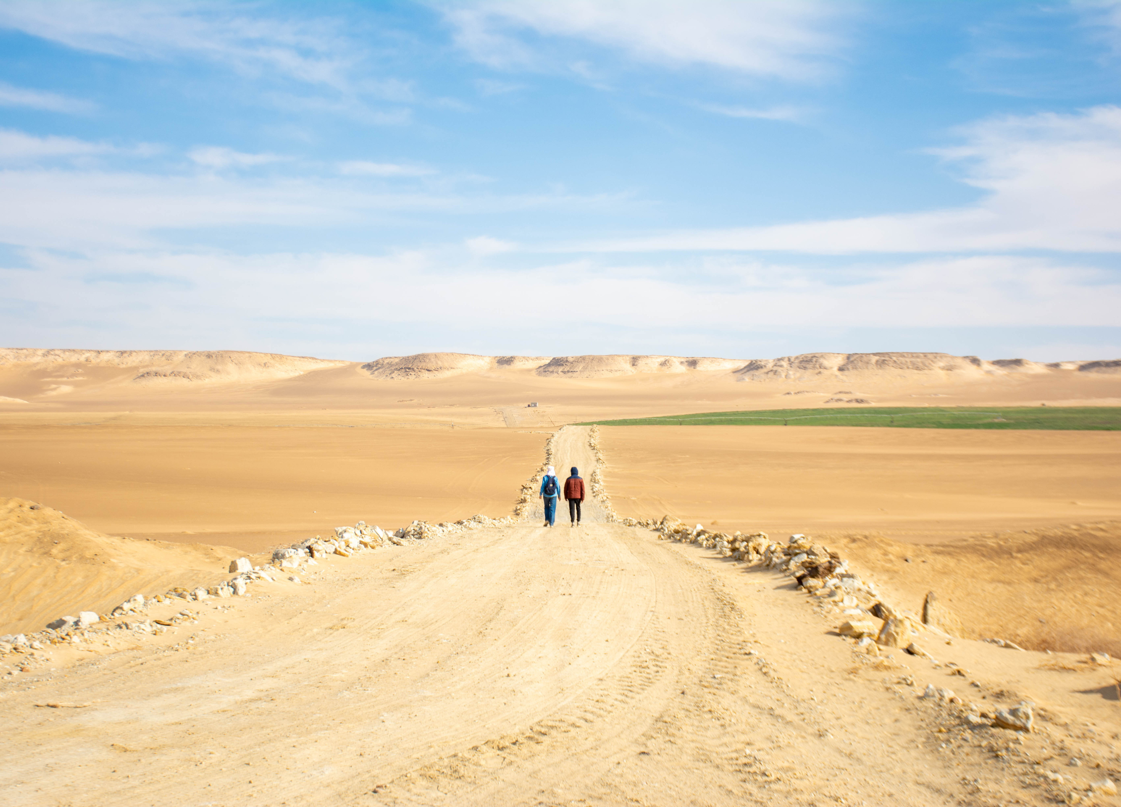 Zwei Menschen gehen auf einer breiten Sandpiste durch eine weite Wüstenlandschaft