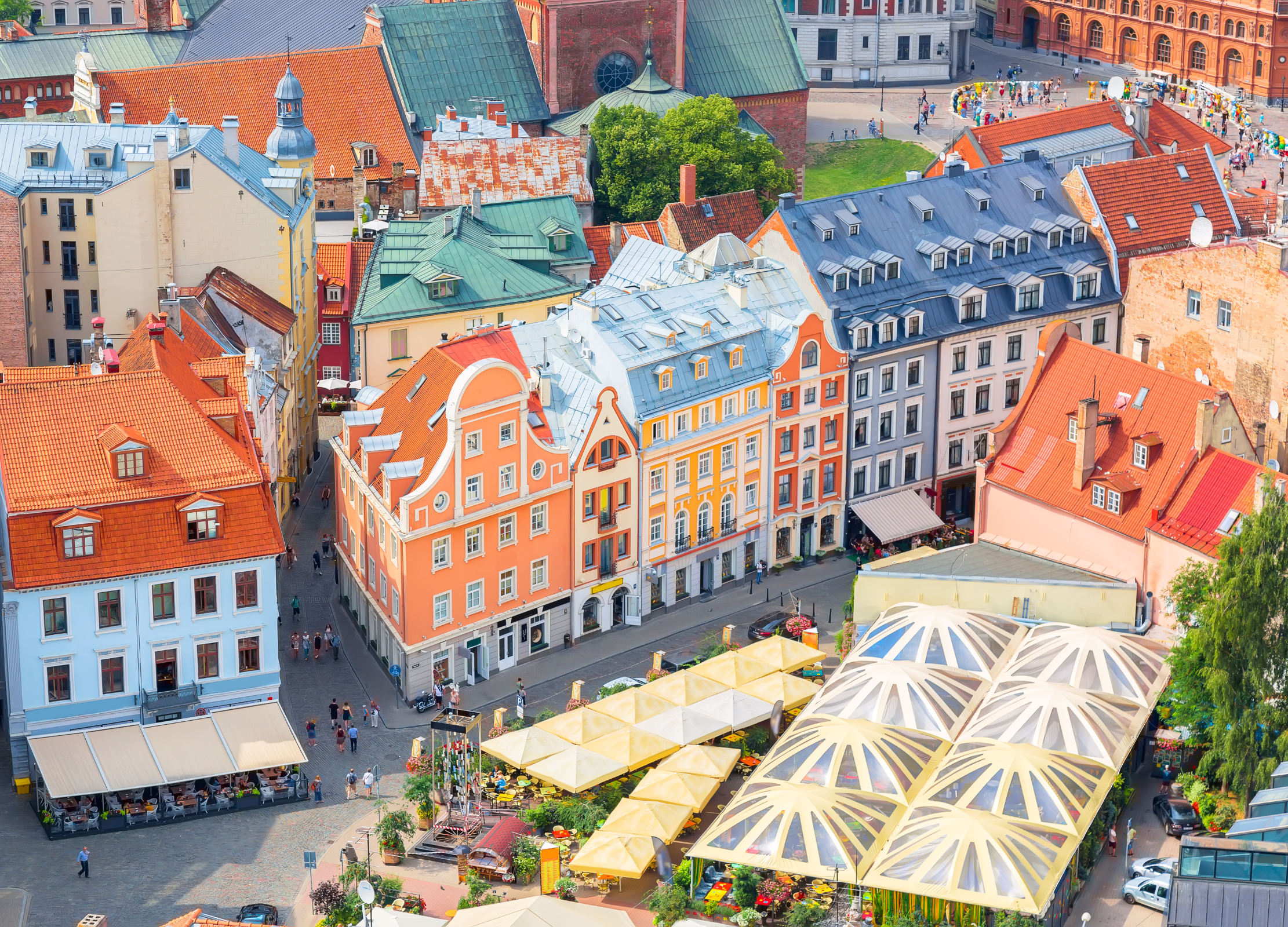 Blick von oben auf einen farbenfrohen historischen Marktplatz mit alten Häusern und Sonnenschirmen, vermutlich in einer lettländischen Altstadt