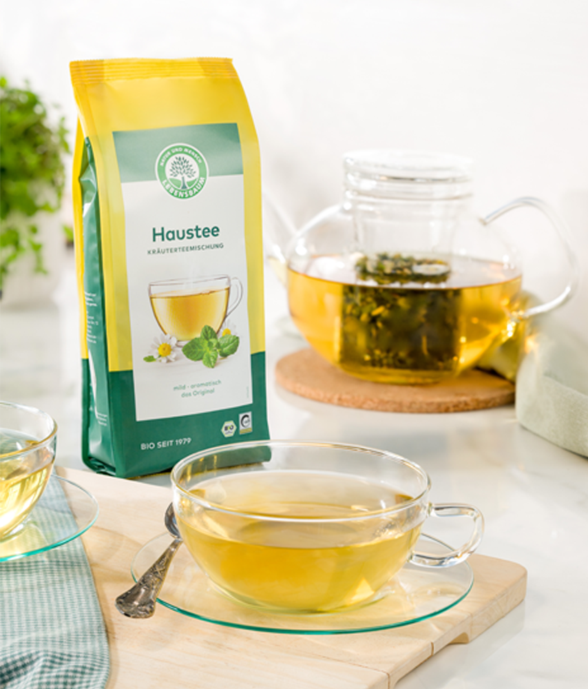 Packung Lebensbaum Haustee steht neben einer Teekanne mit aufgebrühtem Kräutertee und zwei Glastassen auf einem hellen Tisch