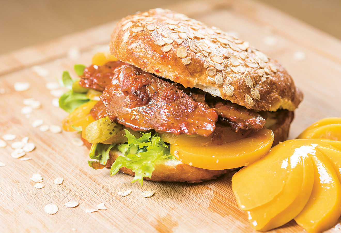 Barbecue-Seitan-Burger auf einem Holzbrett, davor Pfirsichscheiben
