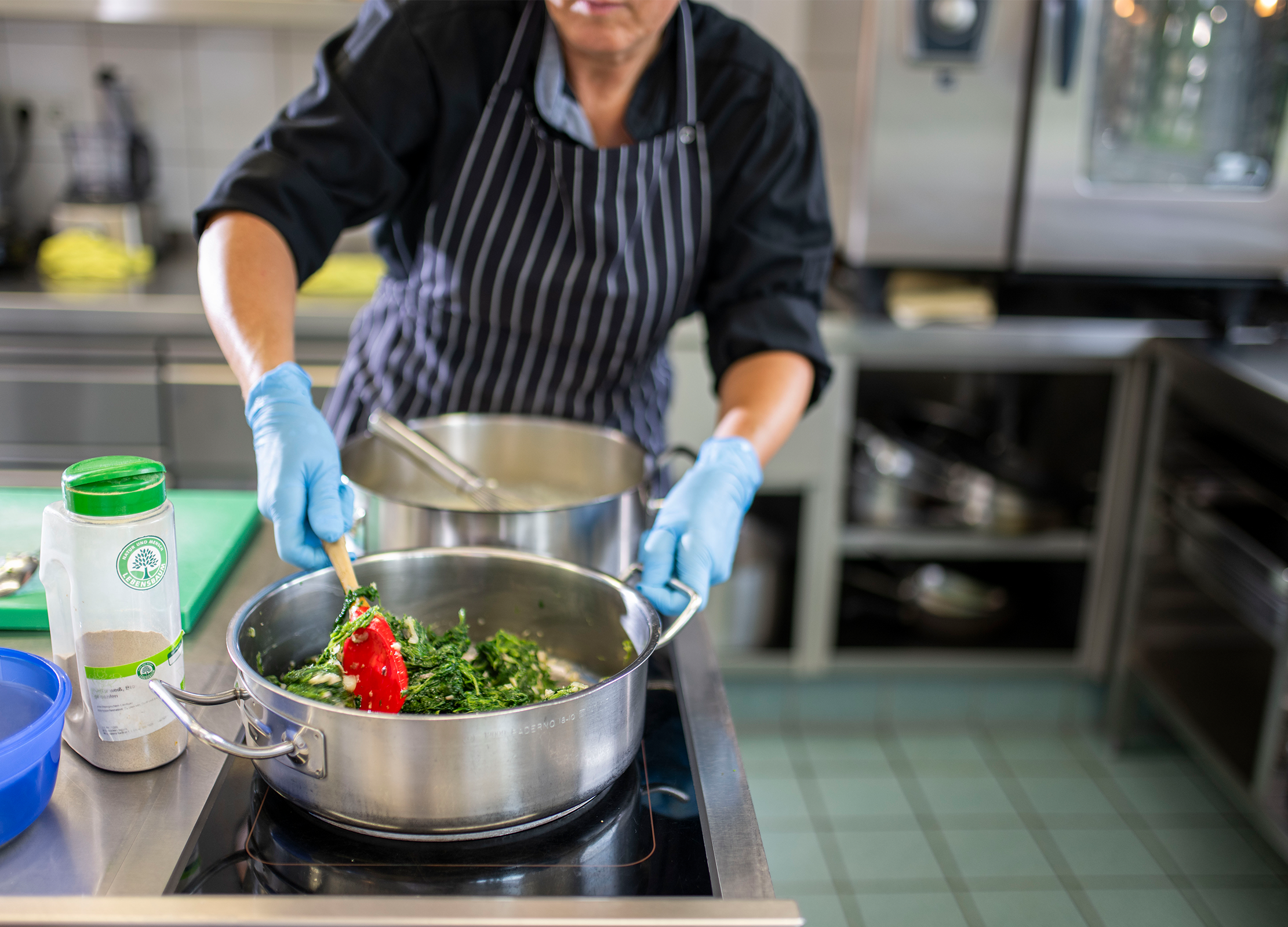 Person in Kochkleidung rührt mit einem roten Löffel Gemüse in einem großen Topf auf dem Herd