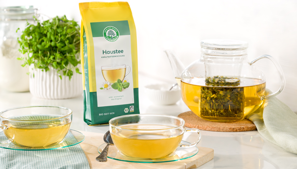 Packung Lebensbaum Haustee steht neben einer Teekanne mit aufgebrühtem Kräutertee und zwei Glastassen auf einem hellen Tisch
