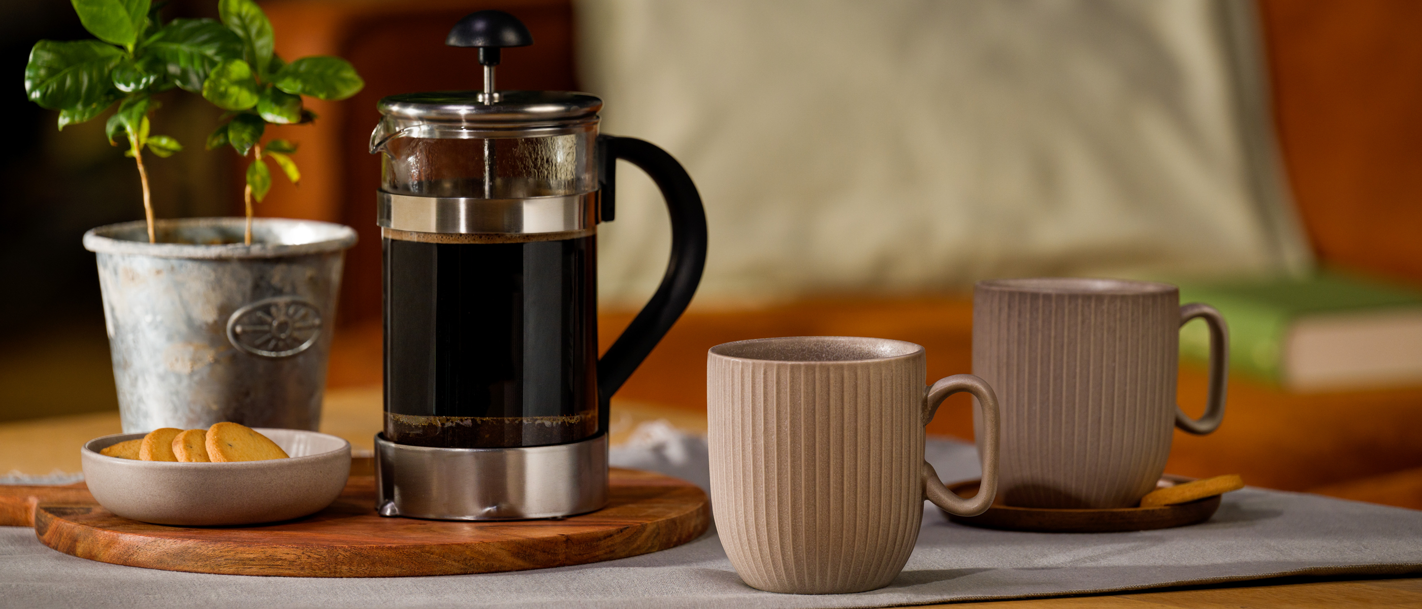 Zwei beige Tassen stehen neben einer gefüllten French Press auf einem gedeckten Tisch, daneben ein kleiner Keksteller und eine Topfpflanze