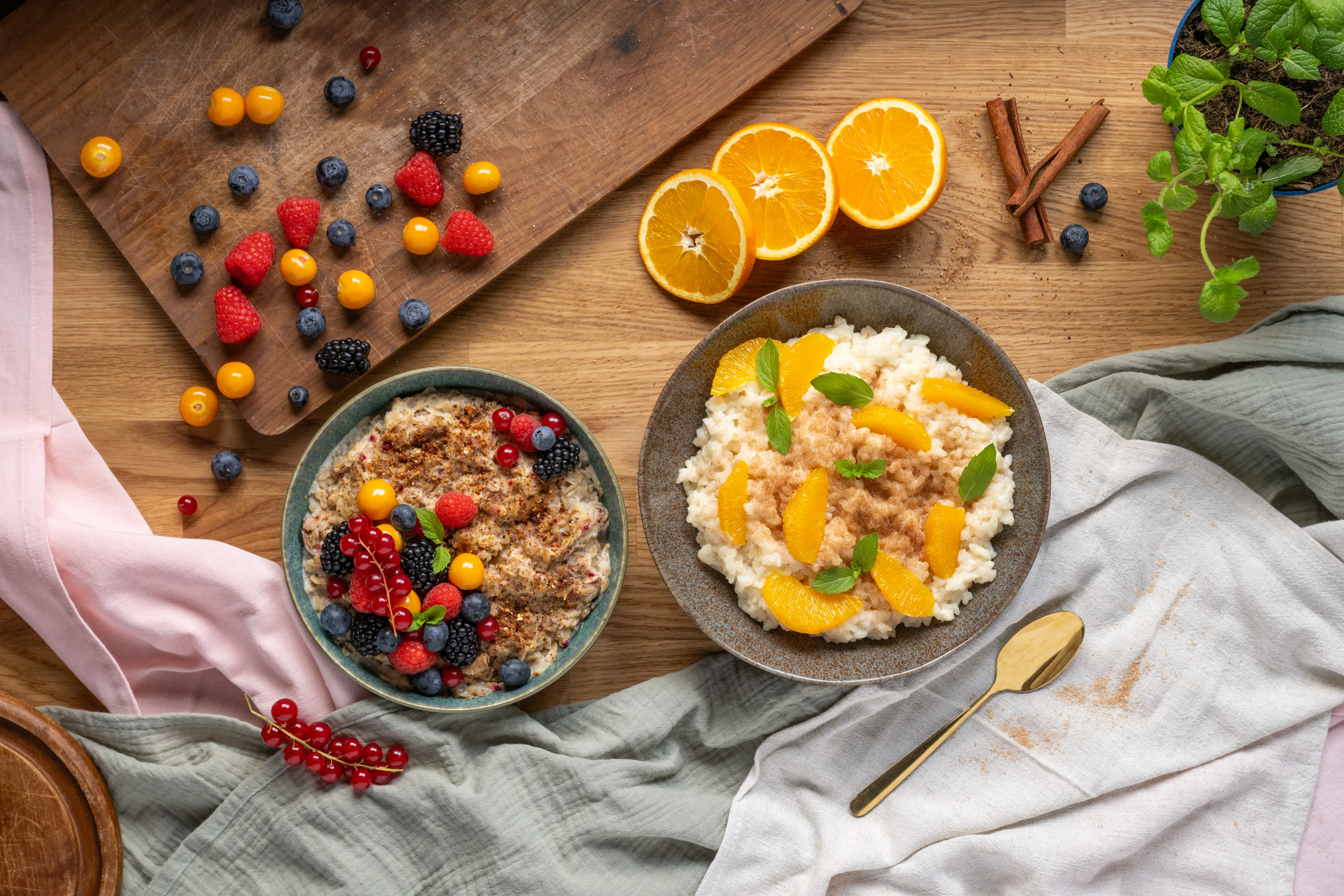 Zwei Frühstücksbowls mit Haferbrei, frischen Beeren und Orangenstücken, umgeben von Zimtstangen, Orangenscheiben und Minze auf Holztisch.