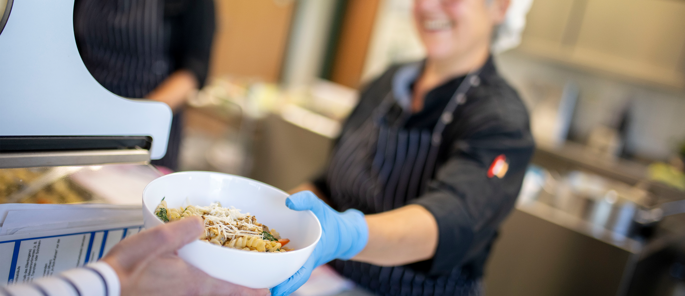 Küchenmitarbeiterin reicht eine Schale mit Pasta über die Essensausgabe