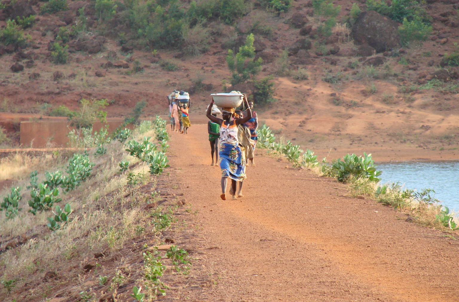 Menschen gehen auf einem roten Weg am Wasser entlang, eine Frau trägt einen Behälter auf dem Kopf in Burkina Faso