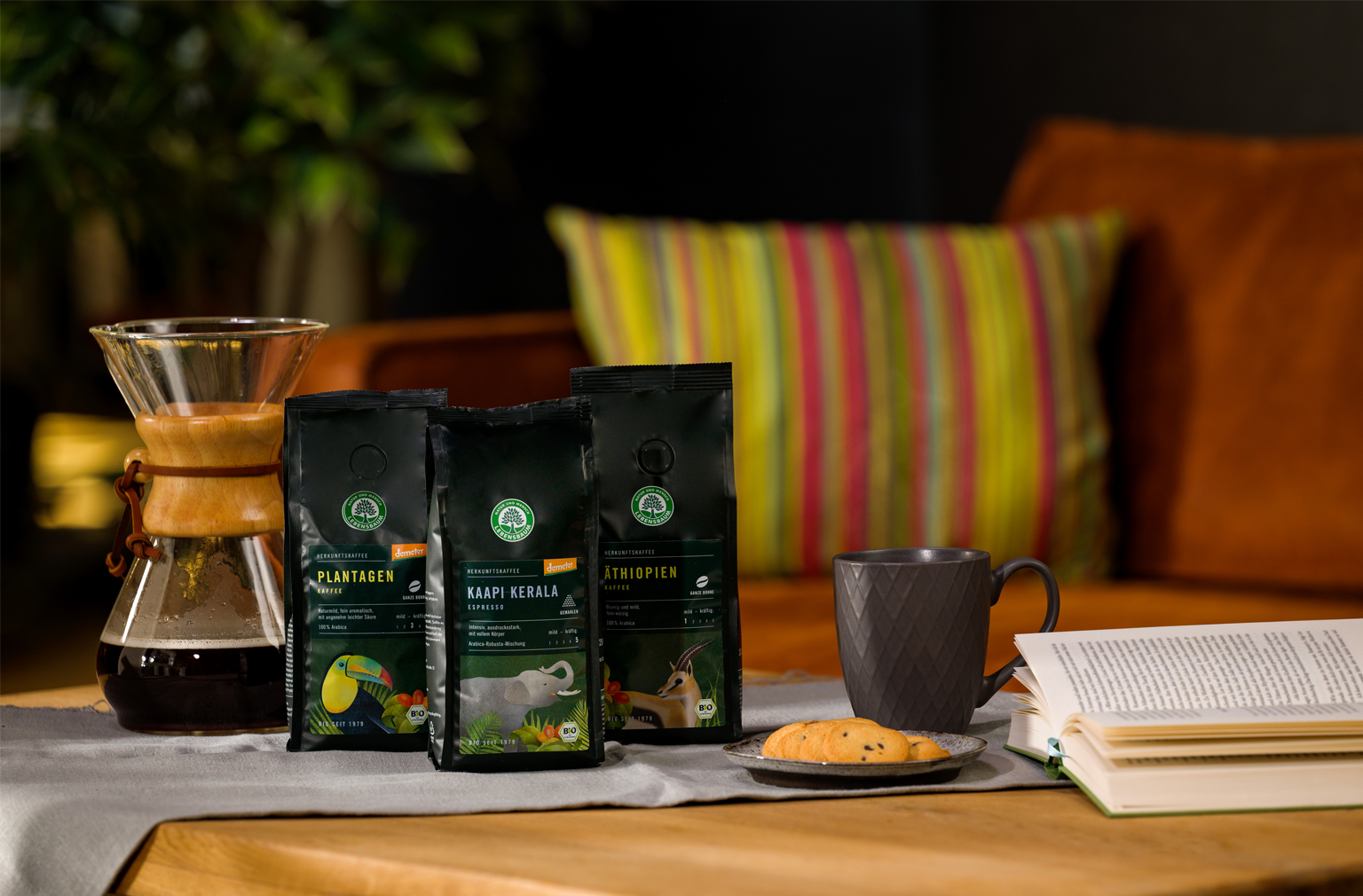 Drei Packungen Lebensbaum-Kaffee stehen auf einem Tisch mit grauer Tasse, Keks und aufgeschlagenem Buch – daneben eine Chemex-Karaffe mit frisch gebrühtem Kaffee