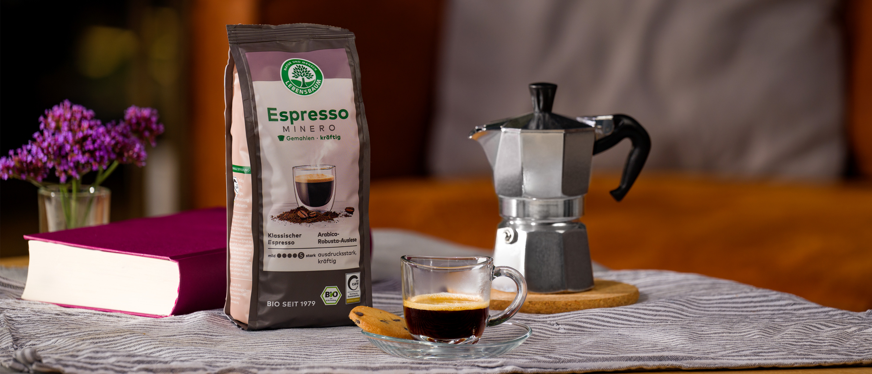 Packung Lebensbaum Espresso Mild steht neben einer Espressokanne und einer Tasse frisch gebrühten Espressos auf einem gedeckten Tisch