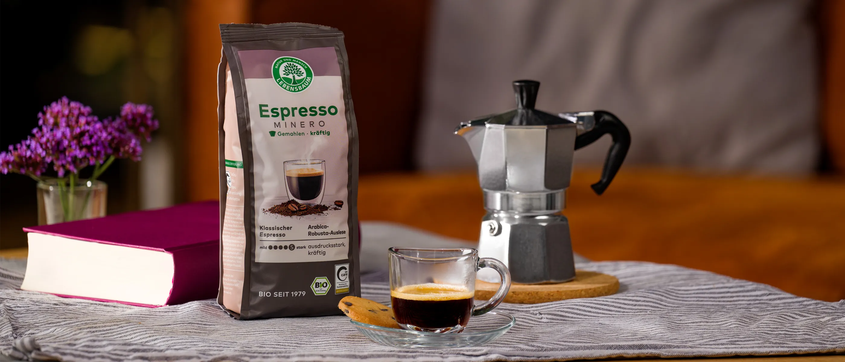 Packung Lebensbaum Espresso Mild steht neben einer Espressokanne und einer Tasse frisch gebrühten Espressos auf einem gedeckten Tisch