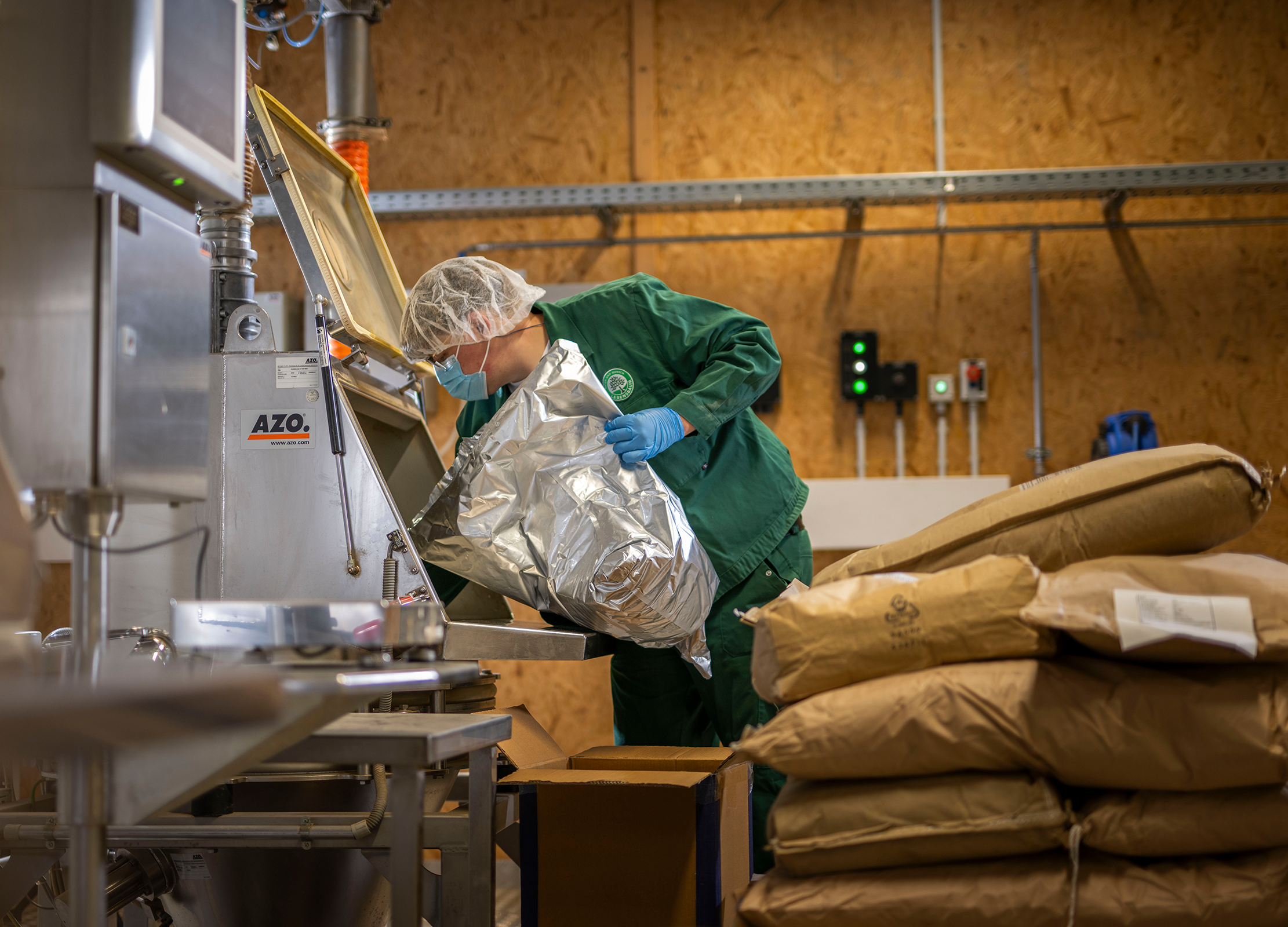 Mischereimitarbeiter in grüner Arbeitskleidung schüttet Kräuter aus einem silbernen Beutel in eine Mischmaschine