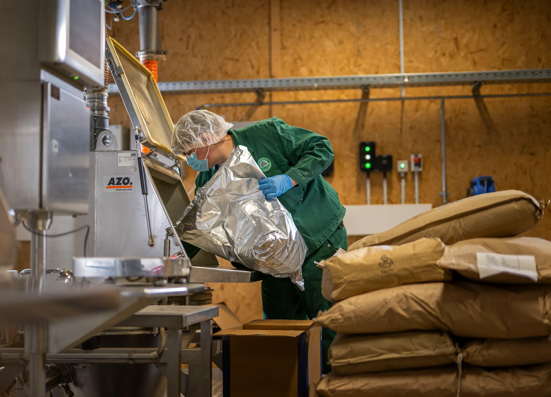Mischereimitarbeiter in grüner Arbeitskleidung schüttet Kräuter aus einem silbernen Beutel in eine Mischmaschine